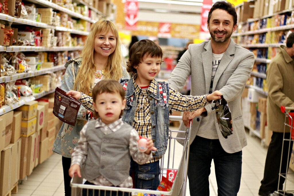 Wife and husband pushing a trolley down a supermarket aisle with their two young children in the trolley.